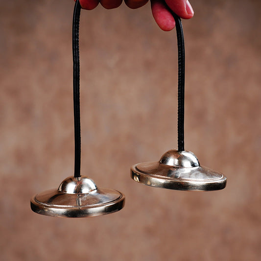 Brassleaf – Smooth polished brass touch bell pair hanging from black cords, held in fingertips against a tan backdrop