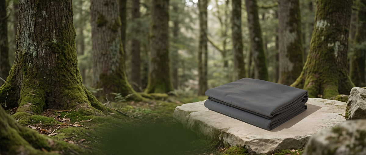 A folded charcoal-grey cotton meditation blanket on a pale stone plinth in a mossy forest.