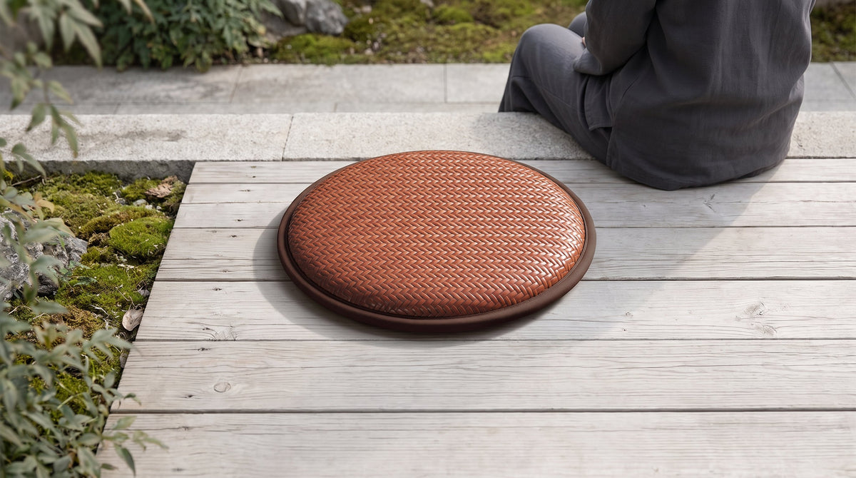 A round warm-brown woven seat cushion on pale timber decking beside moss and stone.