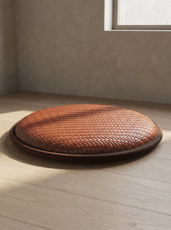 A round woven meditation cushion resting on a pale wood floor in a sunlit plaster room.