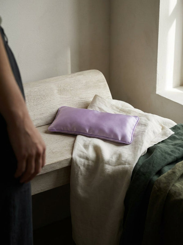 A lavender flaxseed eye pillow resting on folded ivory linen in a quiet bedroom.