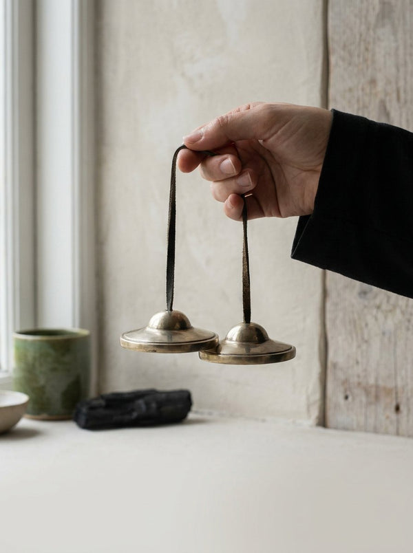 A pair of small copper bells hanging from dark cords in a quiet pale studio.